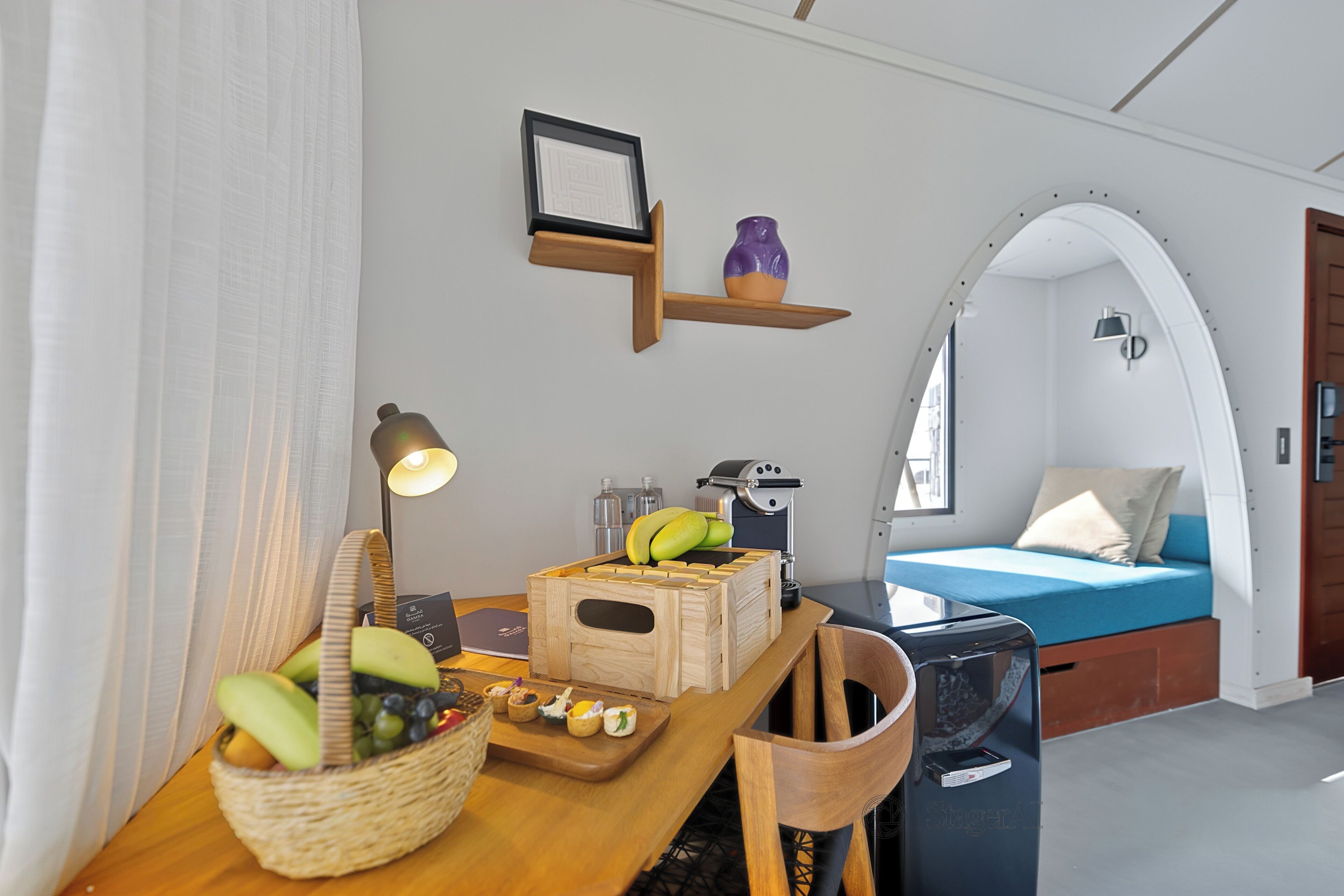 Una habitación moderna con una mesa de madera, una cesta de fruta, una cafetera y una cama con colchón azul.