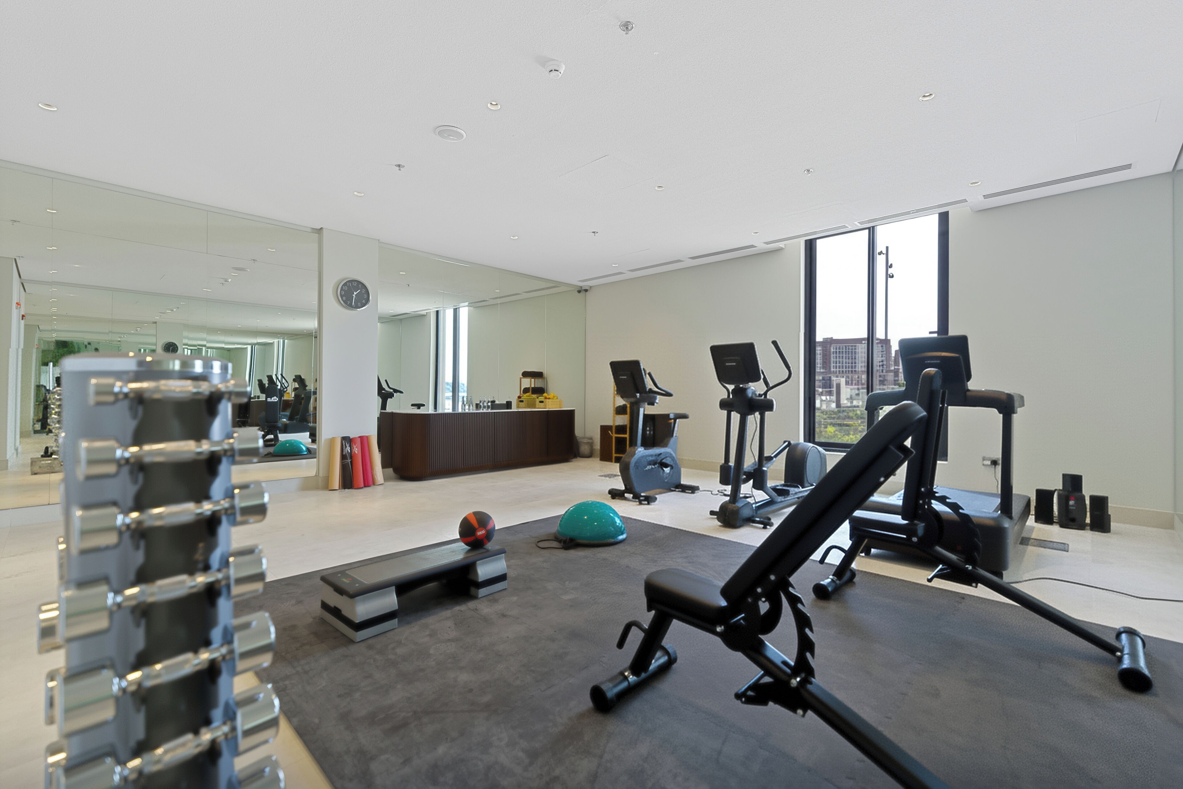Une salle de sport moderne, dotée de divers appareils d’exercice, d’un grand miroir et offrant une vue sur la ville grâce à de larges fenêtres.