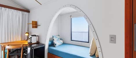 Una habitación compacta con una cama curvada, un escritorio, una silla, una mesita y un ventanal.