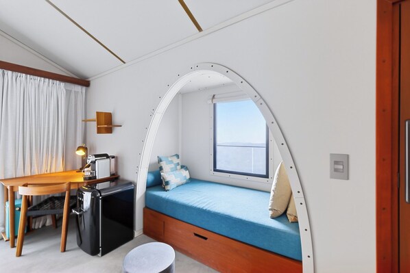 A compact room with a curved bed, a desk, a chair, a small table, and a window.