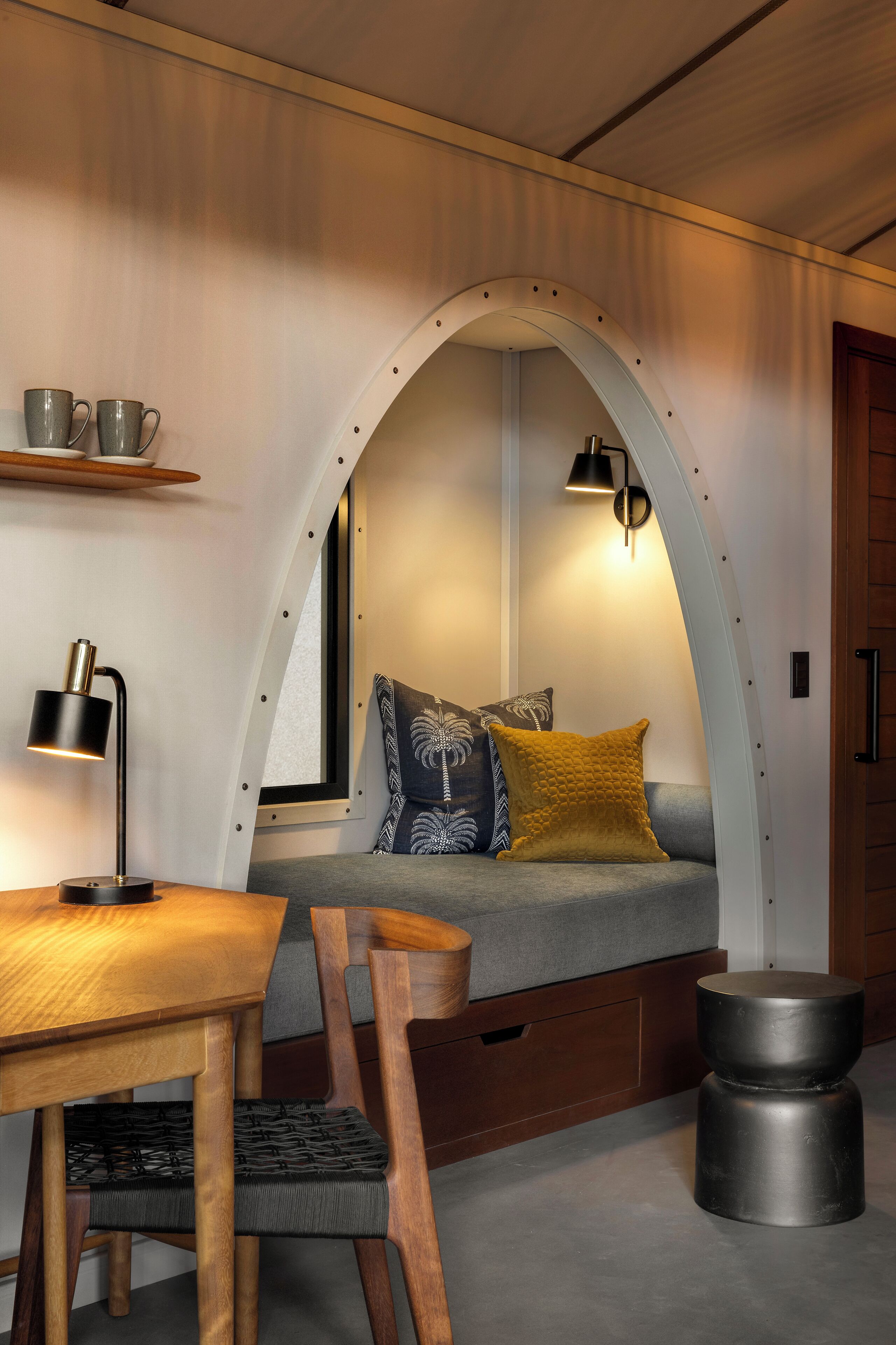 A cozy room with a wooden table, a bench with cushions, a lamp, and a window with a view.