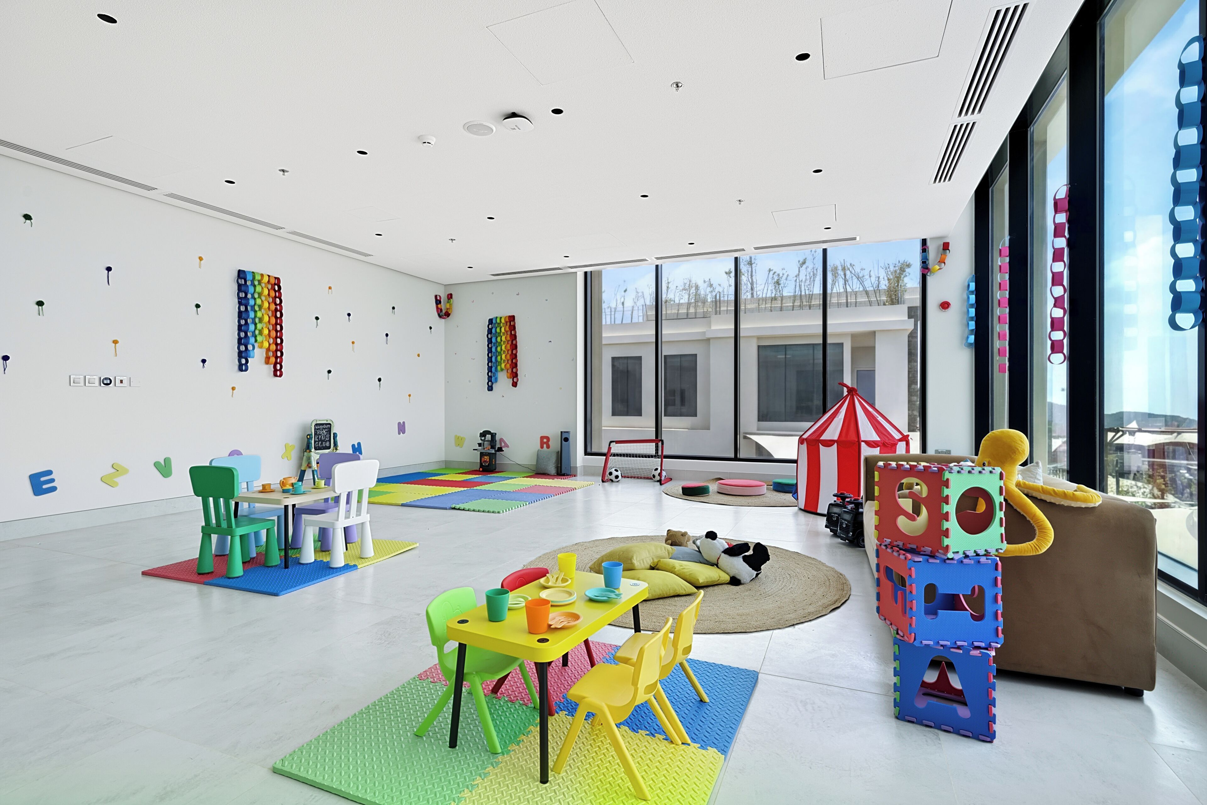 Habitación amplia y luminosa, con un gran ventanal, alfombras de juego coloridas y diversos juguetes y muebles para niños.