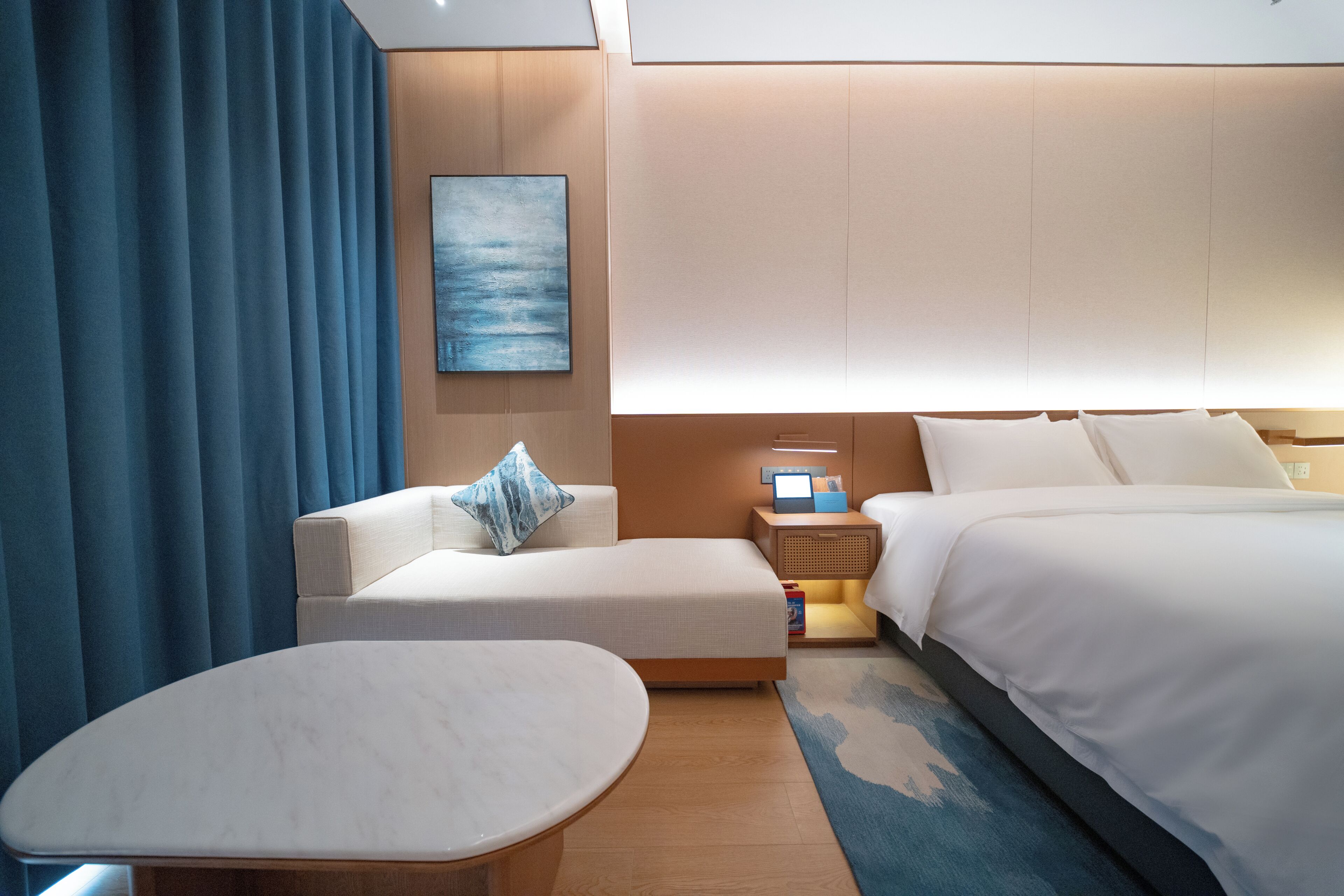A modern hotel room with a bed, a sofa, a small table, a nightstand with a lamp, and a painting on the wall.