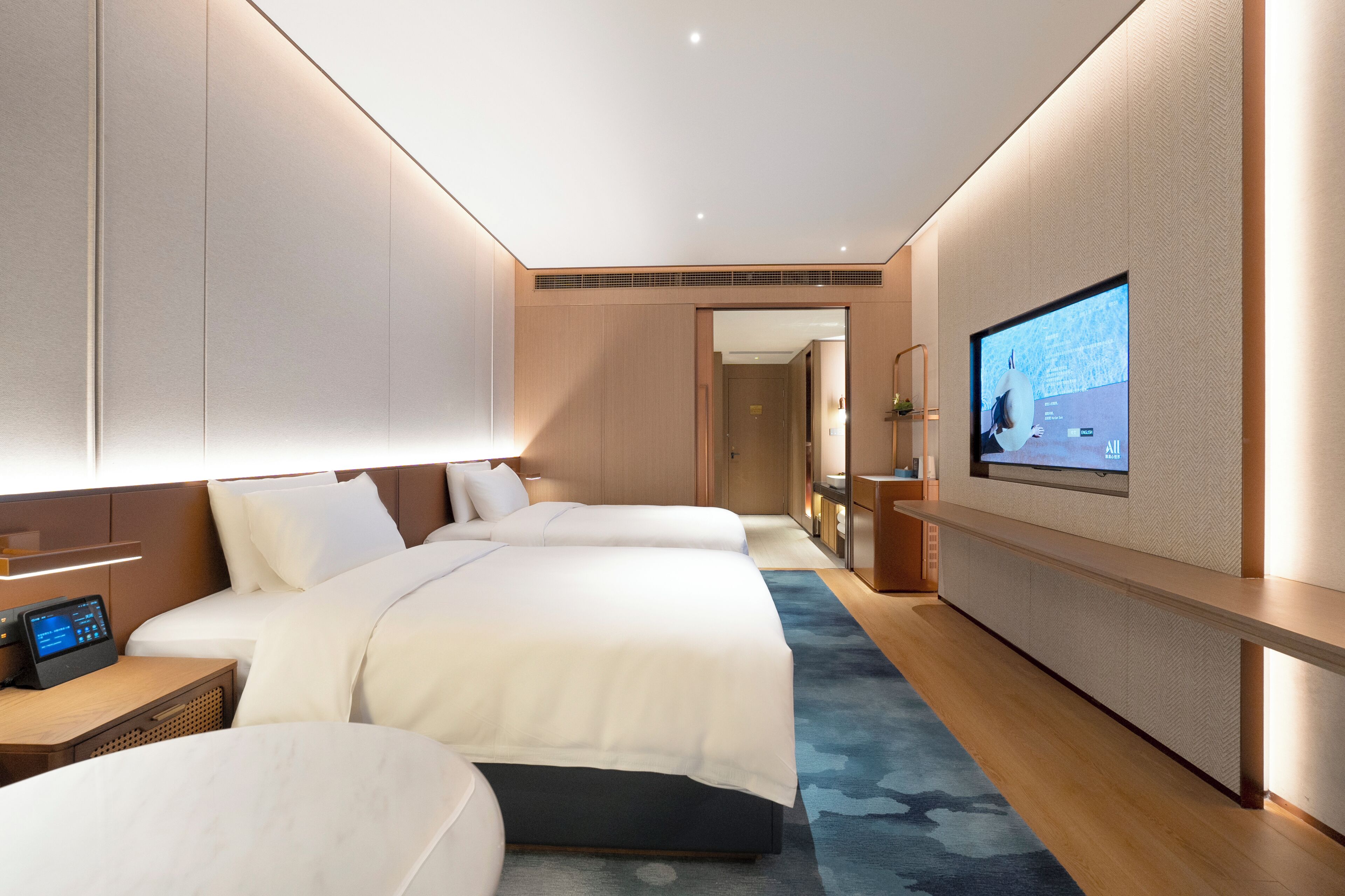 A modern hotel room with two beds, a television, a desk with a computer, and a large window.