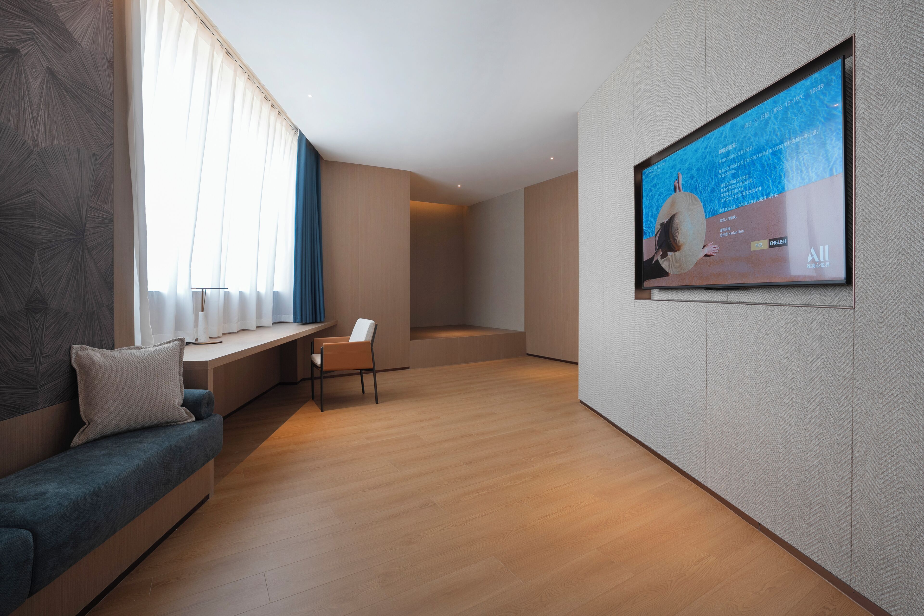 A modern living room with a sofa, a chair, a TV mounted on the wall, and a large window with sheer curtains.