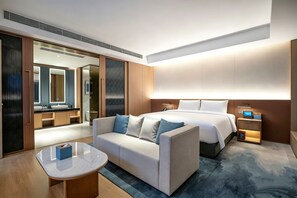 A modern hotel room with a bed, a sofa, a bedside table, a nightstand, and a bathroom visible through an open door. - Grand Mercure Rizhao Haiqu (Rizhao)