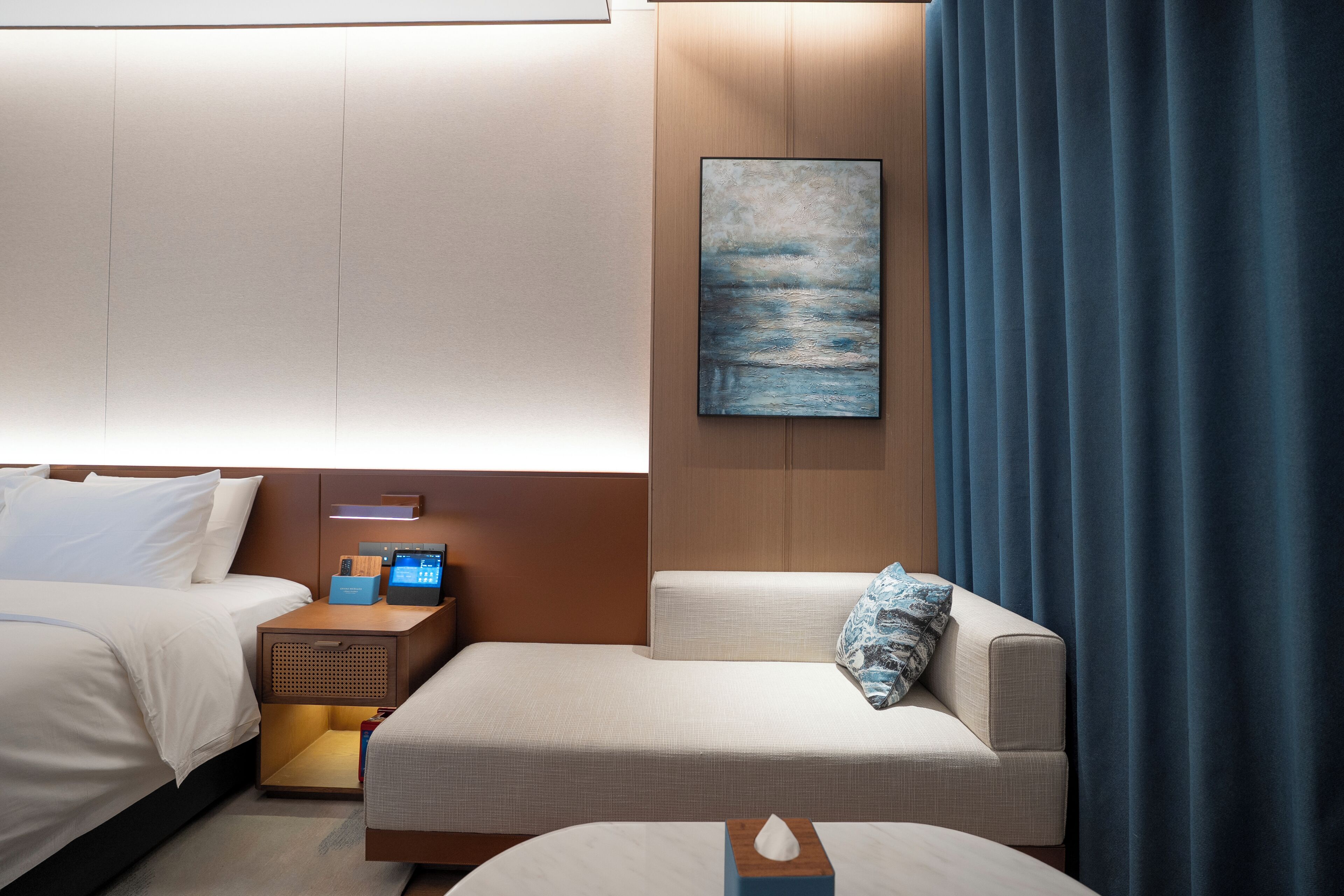 A hotel room with two beds, a bedside table with a lamp, a painting on the wall, and a blue curtain.