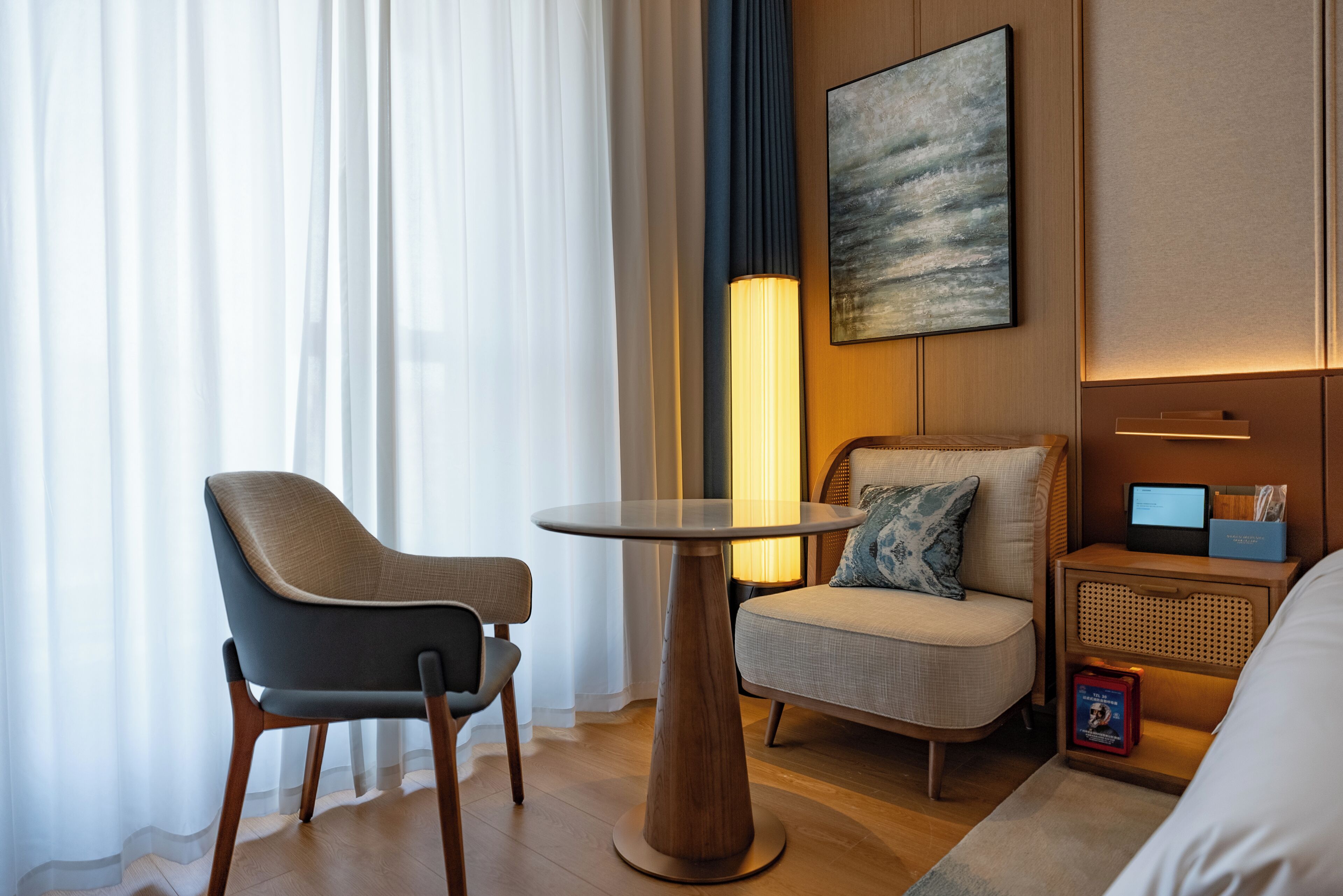 A hotel room with a round table, two chairs, a sofa, a bedside table with a lamp, a wall-mounted artwork, and a small bedside table with a book and a box.
