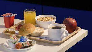 A tray with breakfast items including a muffin, a glass of orange juice, a bowl of cereal, a cup of coffee, a red apple, and a red cup. - Ibis Budget Berlin Ost (Berlin)