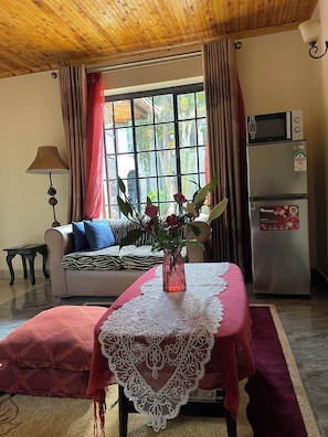 En stue med en sofa, et bord med hvide dug, en vase med blomster, en lampe, et vindue med gardiner og et køleskab.
