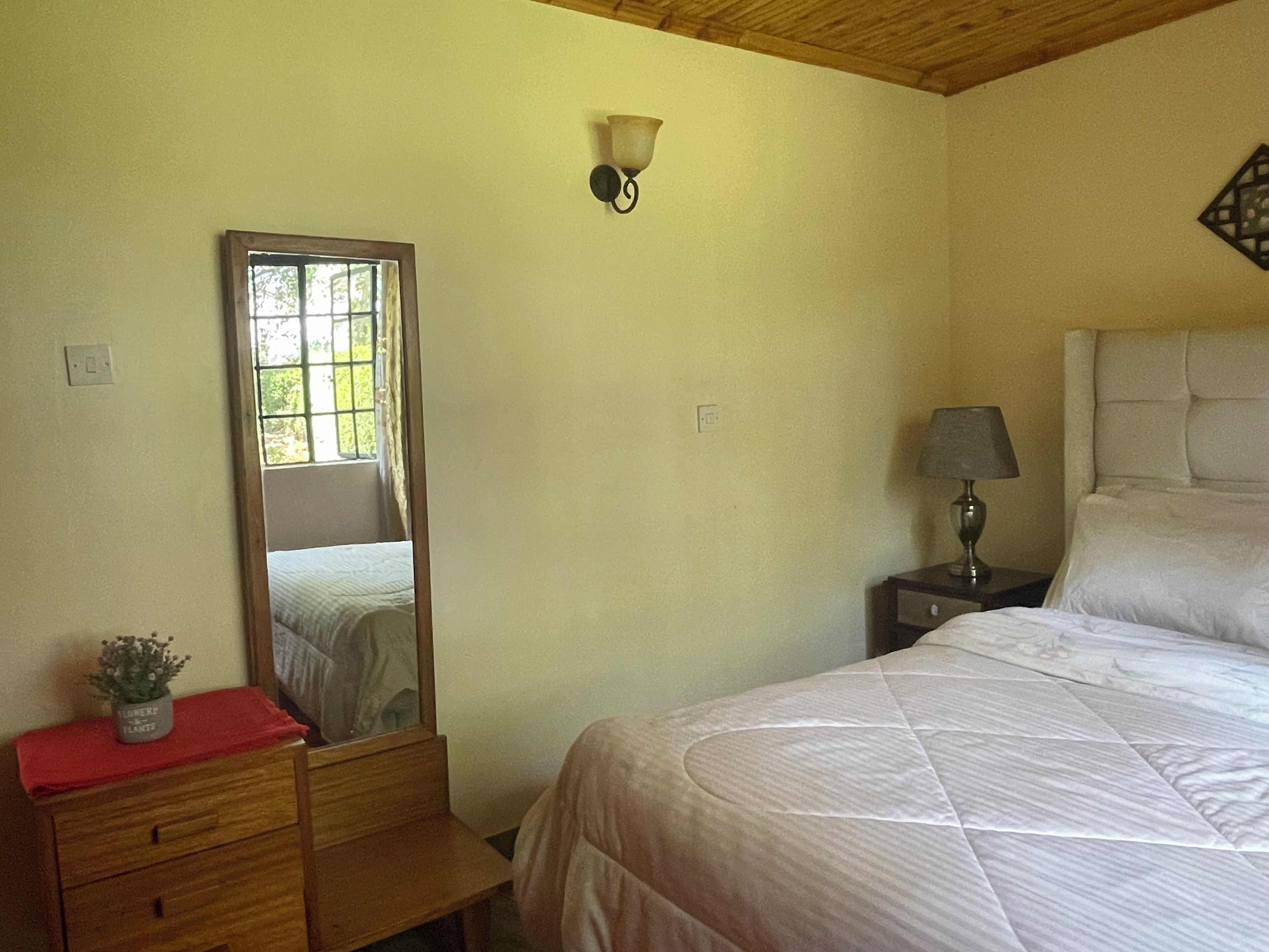 A bedroom with a bed, a nightstand with a lamp, a dresser, a mirror, and a wall-mounted light fixture.