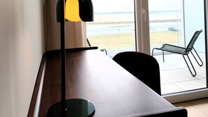 Una habitación con un escritorio de madera, una silla negra, una lámpara de pie y un ventanal corredero con vista a la playa.