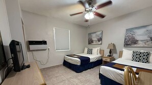 Ein Hotelzimmer mit zwei Betten, einem Schreibtisch, einem Deckenventilator, einem Fernseher, einer Klimaanlage und einem Fenster mit Jalousien.