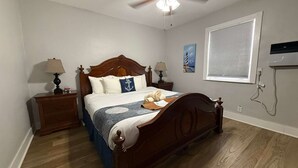 Un dormitorio con una cama de madera, dos mesitas de noche con lámparas, un ventilador de techo, una ventana con persianas y un cuadro en la pared.