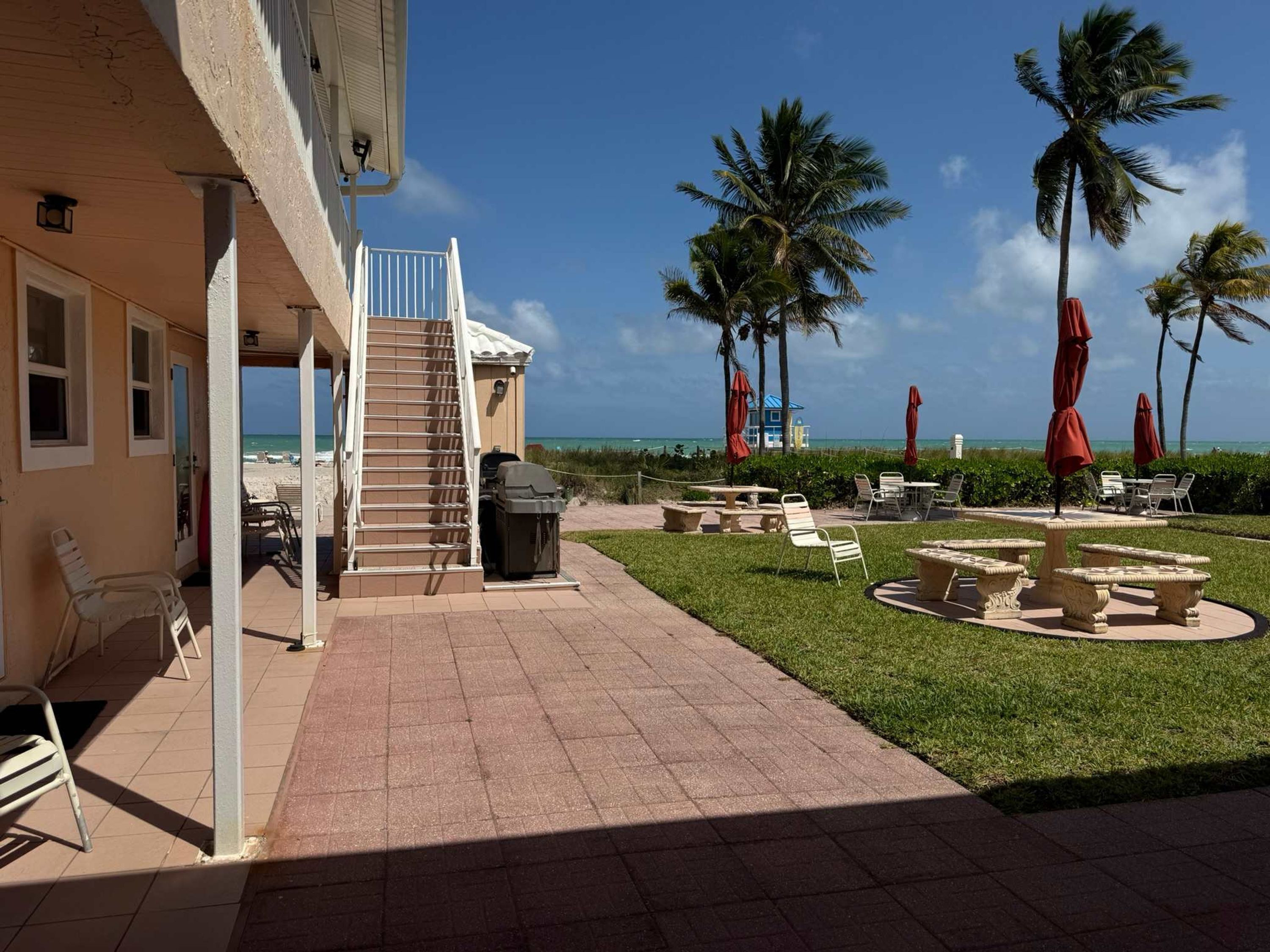 A hotel patio with a staircase leading to a balcony, outdoor seating, palm trees, and a view of the beach.