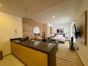 Quarto de hotel moderno com cozinha, cama e televisão grande, e piso de madeira.