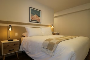 Ein ordentlich bezogenes Bett mit einer karierten Tagesdecke, zwei Nachttischlampen, einem Nachttisch mit Schublade und einem gerahmten Bild an der Wand.