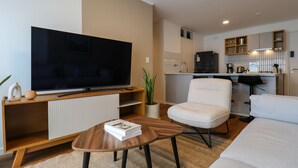 Una sala de estar moderna con un televisor de pantalla plana, un sofá blanco, una mesa de centro y una cocina al fondo.