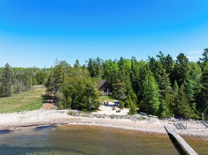 Una cabaña rodeada de un bosque frondoso, una playa de arena y un muelle de madera que se adentra en un río.