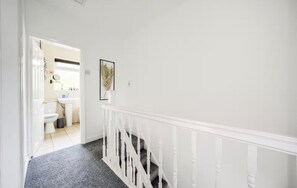 A hallway with a white railing, a staircase, and a bathroom visible in the background.