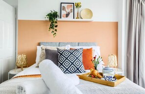 A neatly made bed with a variety of pillows, a bedside table with a tray of food and a teapot, and a wall-mounted shelf with decorative items.