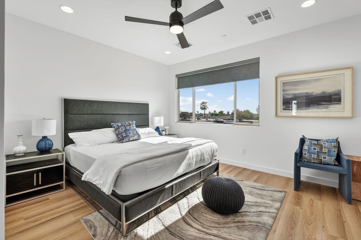 A modern bedroom with a bed, nightstands, a chair, a ceiling fan, and a window with a view.