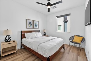 Een slaapkamer met een bed, twee nachtkastjes, een stoel, een plafondventilator en kunst aan de muur.