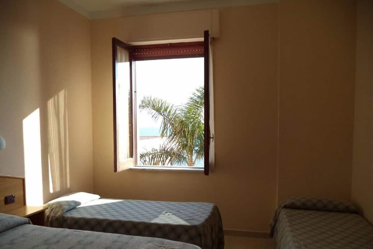 A room with two beds, a window with a view of palm trees, and a door.