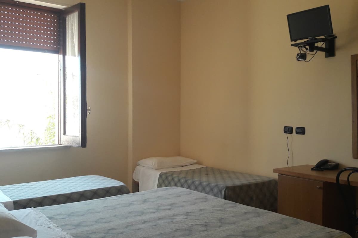 A room with two beds, a desk with a chair, a television mounted on the wall, and a window with blinds.