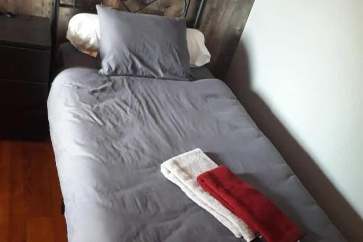 Una cama individual con colchón gris y estructura metálica, una almohada blanca, una almohada gris y una manta roja.