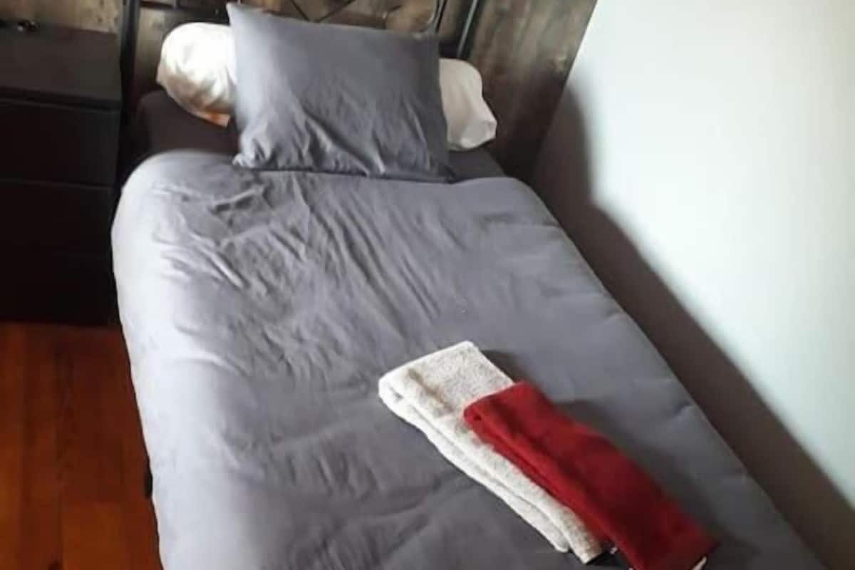 Una cama individual con colchón gris, cabecera de metal y manta roja.