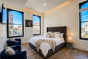 Ein Schlafzimmer mit einem großen Bett, zwei Fenstern, einem Sessel und einem Nachttisch.