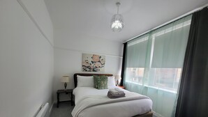 A bedroom with a bed, bedside tables, a hanging light fixture, a window with curtains, and a wall-mounted artwork. - Palace Court in the Heart of Buxton (Buxton)