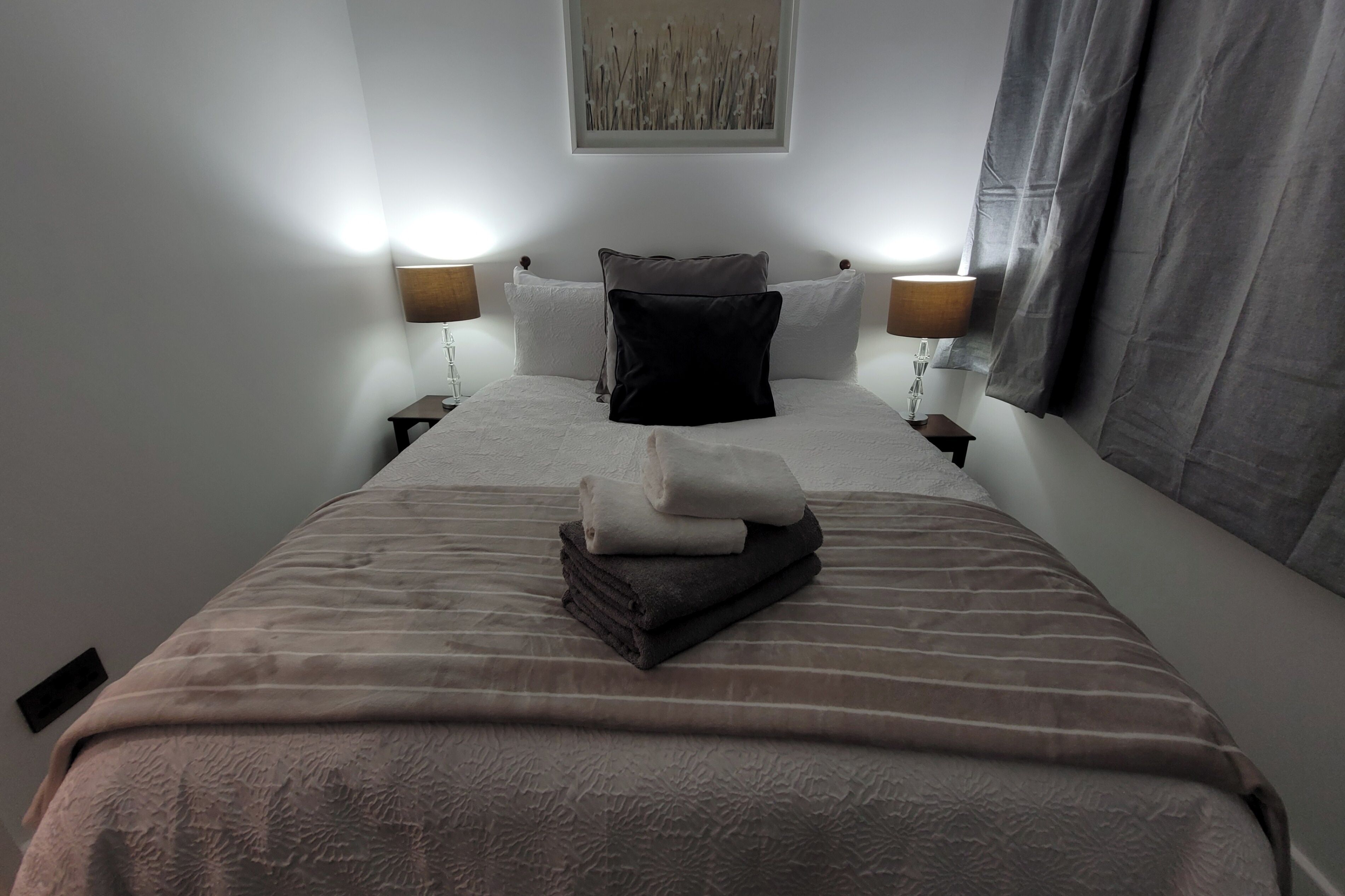 A hotel room with a neatly made bed, two bedside lamps, a wall-mounted artwork, and a window with curtains.