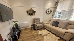 A living room with a sofa, armchair, television, wall clocks, and a radiator. - Palace Court in the Heart of Buxton (Buxton)