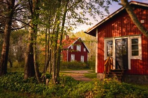 Une maison en bois rouge, dotée d’un porche, entourée d’arbres et accessible par un chemin.