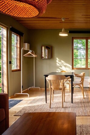 Una habitación con una mesa de madera, sillas, una lámpara colgante, una lámpara de pie y ventanas grandes.