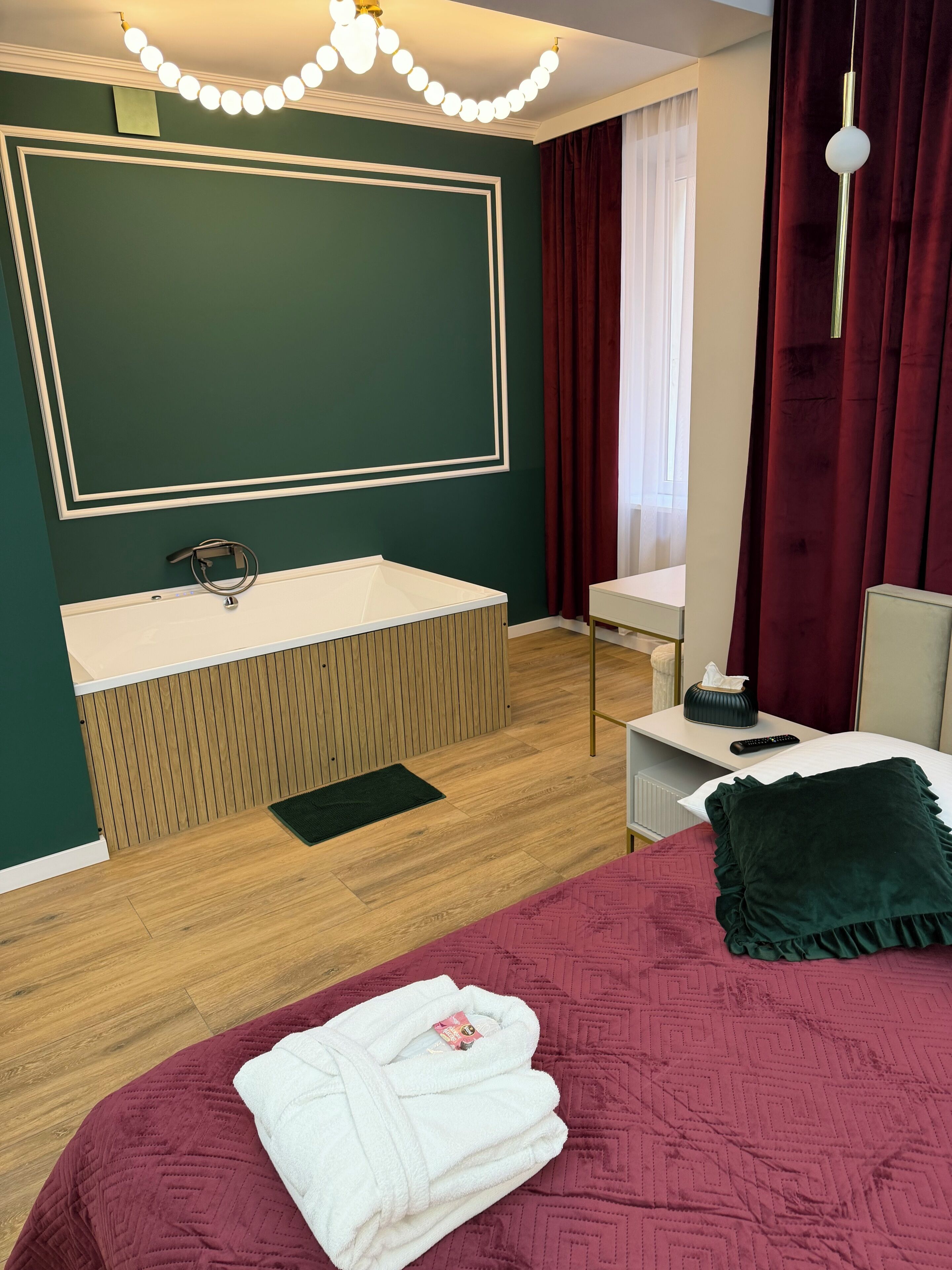 A hotel room with a large bed, a bedside table, a small bedside table, a mirror, a vanity with a sink, and a window with curtains.