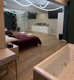 Ein modernes Hotelzimmer mit Holzboden, einem großen Bett mit roter Bettdecke, einem weißen Nachttisch, einem weißen Badezimmer mit Badewanne und einem an der Wand befestigten Fernseher.