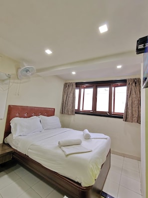 Un lit bien fait, avec du linge de lit blanc, une tĂȘte de lit en bois, un ventilateur de plafond et une fenĂȘtre avec des rideaux.