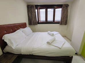 Un lit bien fait, avec du linge de lit et des serviettes blanches, une tĂȘte de lit rembourrĂ©e de couleur marron et une fenĂȘtre avec des rideaux.