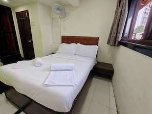 A neatly made bed with white linens and towels, a wooden headboard, a wall-mounted fan, and a window with curtains.