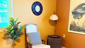 A room with a wooden floor, a chair with a cushion, a potted plant, a wall clock, a lamp, and a framed picture. - Captain’s Quarters- charming studio apartment in the countryside (Berlin Center)