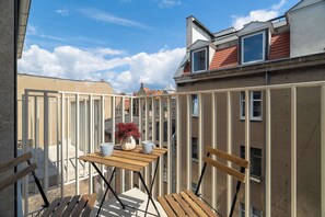 Ein Balkon mit Holztisch, zwei Stühlen, einer Blumenvase und Blick auf angrenzende Gebäude.