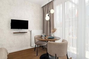 Ein modernes Wohnzimmer mit einem Wand-Fernseher, einem runden Esstisch mit Stühlen und einem großen Fenster mit durchsichtigen Vorhängen.
