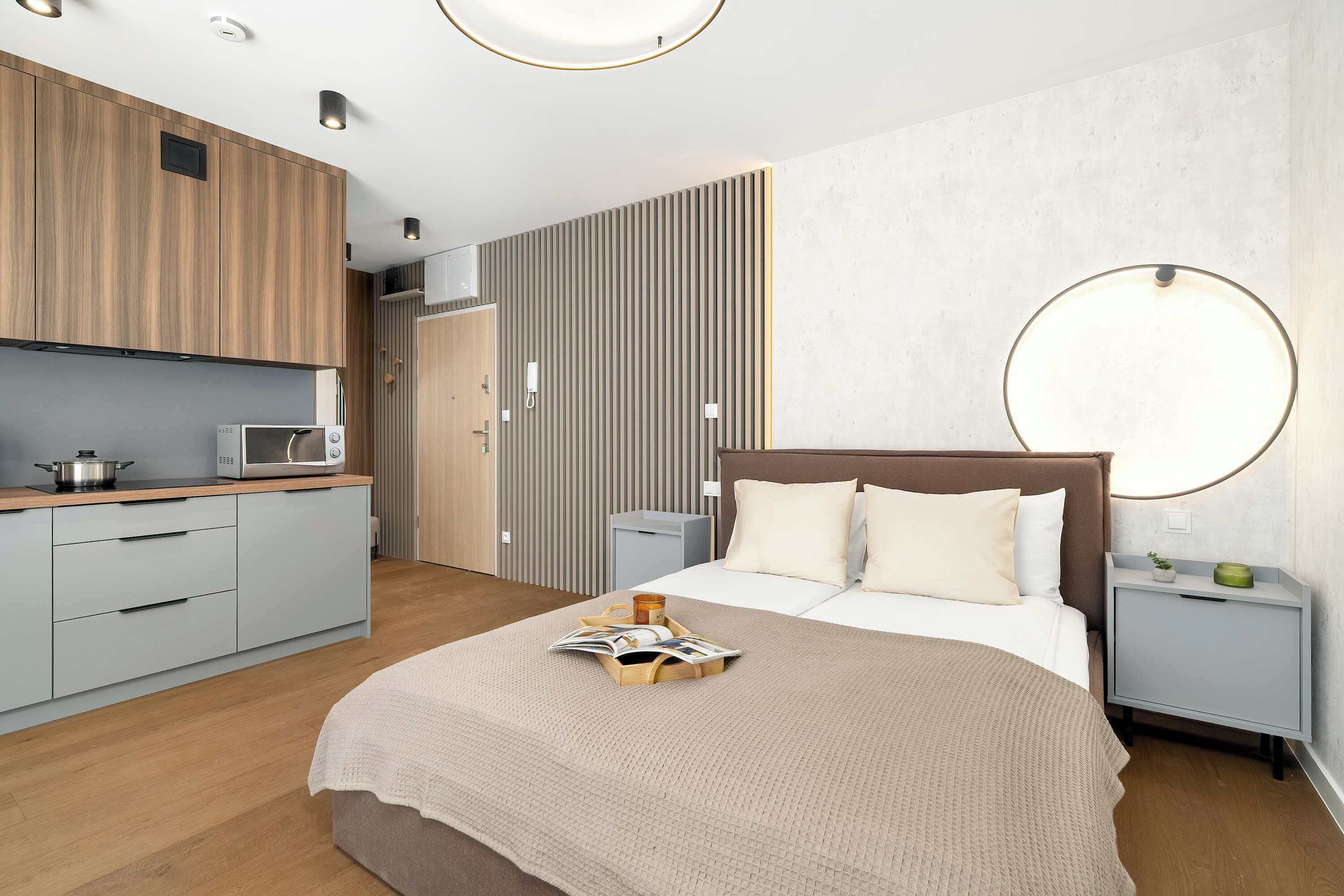 A modern bedroom with a bed, bedside tables, a nightstand, a microwave, and a kitchen area with cabinets and a countertop.