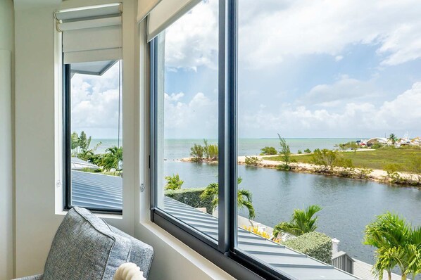 Ein Zimmer mit großen Fenstern und Blick auf ein Gewässer, das Meer sowie eine entfernte Küstenlinie mit Gebäuden und Grünflächen.