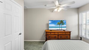 A bedroom with a bed, a wooden dresser, a ceiling fan, a painting of a beach, and a door. - Saltwater Heaven (Rehoboth Beach)