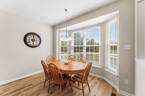 A dining room with a round table, wooden chairs, a wall clock, and large windows with blinds. - Saltwater Heaven (Rehoboth Beach)