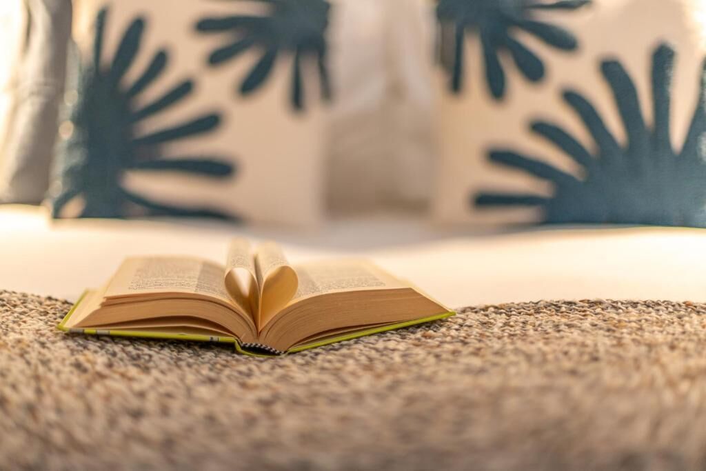 Un livre ouvert, posé sur une surface texturée, vraisemblablement un lit ou un canapé, avec un arrière-plan flou où l’on aperçoit un coussin orné d’un motif de feuilles.